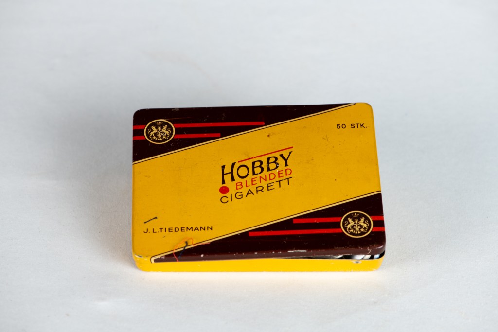 Bilde av en sigarettboks med teksten 'HOBBY BLENDED CIGARETT' i gult og brunt, datert fra midten av 1900-tallet.