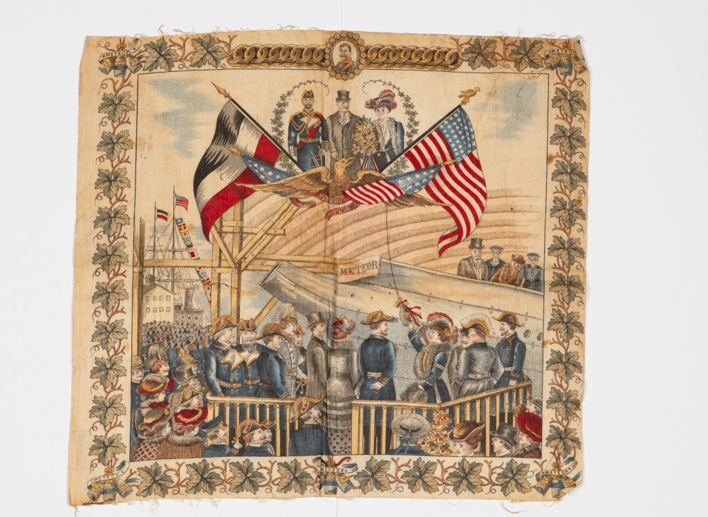 Eit fargerikt bilde som skildrar ei seremoni med fleire personar som heiser flagg, inkludert det amerikanske og tyske flagget, med ei folkemengde til stades. Biletet har også et dekorerande ramme med blomar.