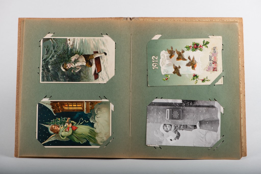 Eit album med gamle julekort og ein fotografi, inkludert motiv av ung gut med tre og engel med julelys.
