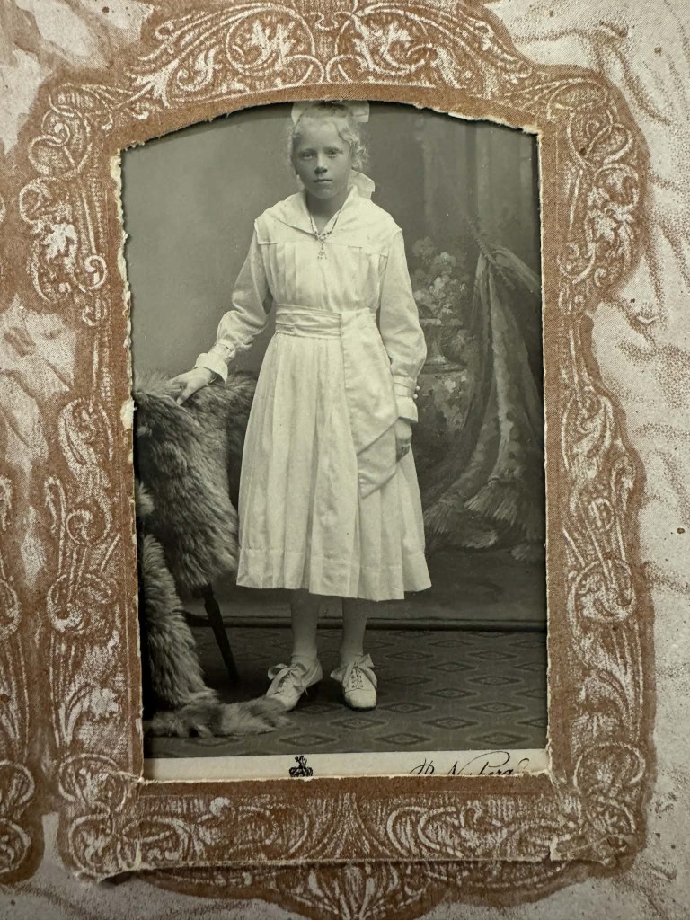 Eit gammalt svart-kvitt fotografi av ei jente i kvit kjole som står ved sidan av eit pelsforetak.